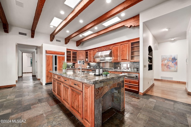 kitchen with a skylight, stone tile floors, an island with sink, wall chimney exhaust hood, and glass insert cabinets