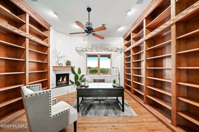 office space featuring a warm lit fireplace, ceiling fan, and wood finished floors