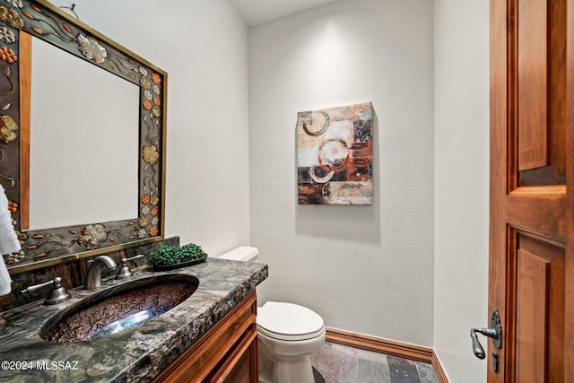 half bathroom with toilet, stone finish floor, vanity, and baseboards