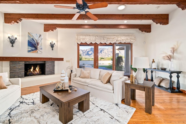 living area featuring light wood-style flooring, beamed ceiling, a stone fireplace, and ceiling fan