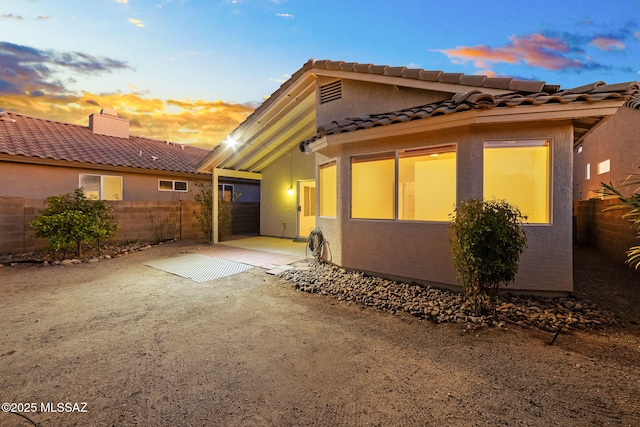 property exterior at dusk with a patio, a tile roof, fence, and stucco siding