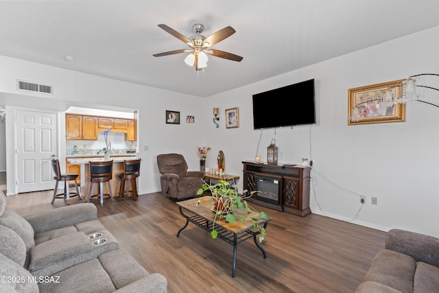 living area with dark wood finished floors, baseboards, visible vents, a ceiling fan, and a glass covered fireplace