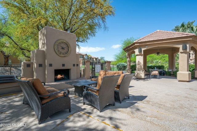 view of patio / terrace with a warm lit fireplace, ceiling fan, fence, and a gazebo