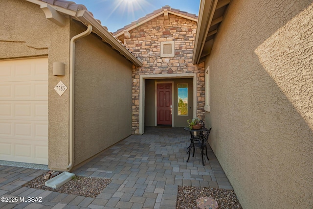 view of exterior entry with an attached garage, stone siding, a tiled roof, and stucco siding