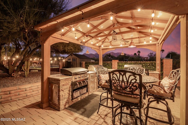 patio terrace at dusk with a gazebo, area for grilling, fence, and a ceiling fan