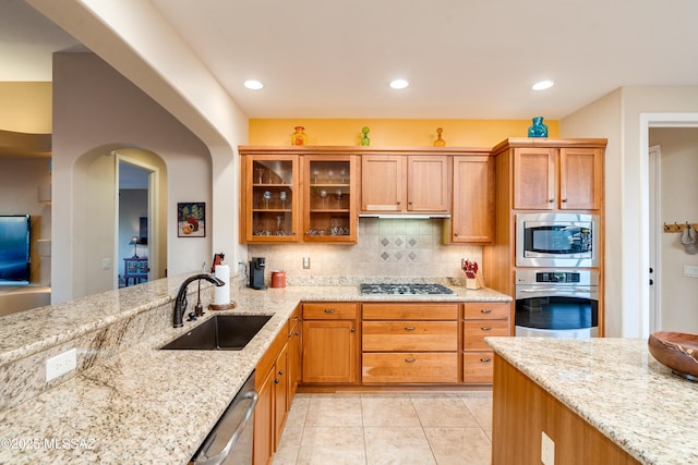 kitchen with light stone counters, a sink, appliances with stainless steel finishes, tasteful backsplash, and glass insert cabinets