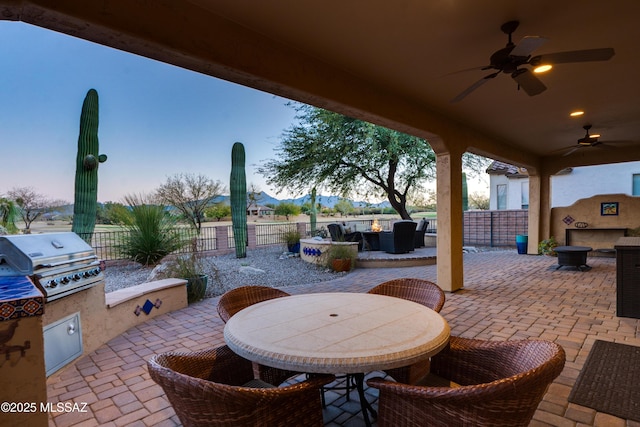 patio terrace at dusk featuring outdoor dining area, area for grilling, fence, a ceiling fan, and exterior kitchen