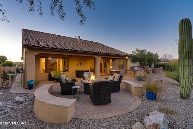 back of property at dusk with an outdoor living space with a fire pit, an outdoor kitchen, a patio area, fence, and a tiled roof