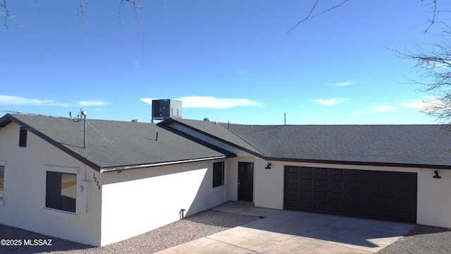 ranch-style house featuring driveway, roof with shingles, an attached garage, central air condition unit, and stucco siding