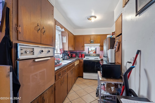 kitchen featuring electric stove, light tile patterned floors, brown cabinetry, freestanding refrigerator, and under cabinet range hood