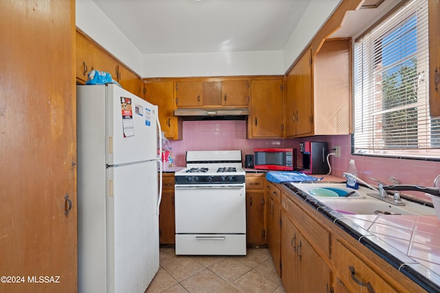 kitchen featuring tile countertops, under cabinet range hood, white appliances, a sink, and brown cabinetry