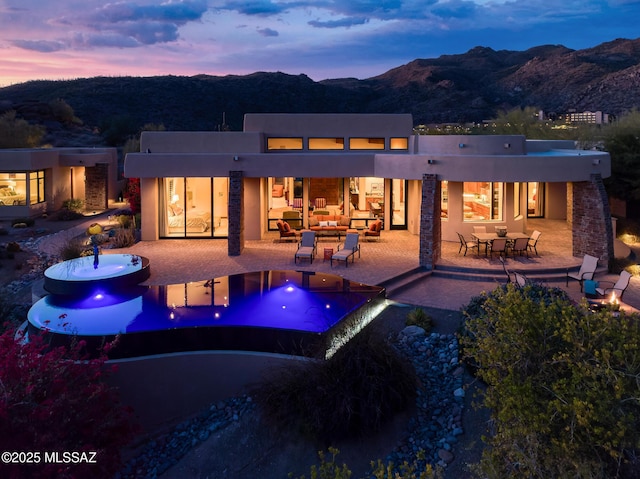 back of property at dusk featuring a patio, stucco siding, outdoor lounge area, a mountain view, and an infinity pool