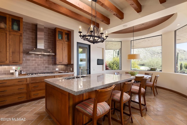 kitchen with a sink, glass insert cabinets, wall chimney exhaust hood, brown cabinetry, and decorative backsplash