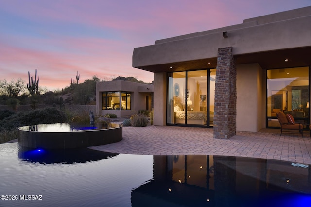 back of property at dusk with a patio, stucco siding, and an infinity pool