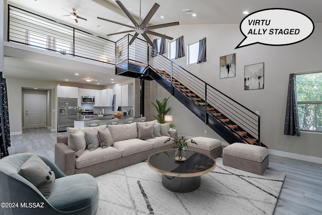 living room featuring light wood-style flooring, a ceiling fan, stairs, and a towering ceiling