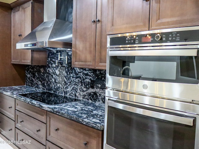 kitchen featuring stainless steel double oven, dark stone counters, black electric stovetop, wall chimney range hood, and backsplash
