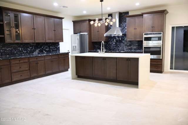 kitchen with an island with sink, a sink, pendant lighting, appliances with stainless steel finishes, and wall chimney range hood