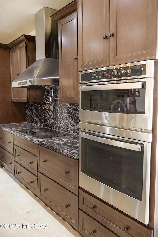 kitchen with dark stone countertops, black electric stovetop, double oven, wall chimney range hood, and backsplash