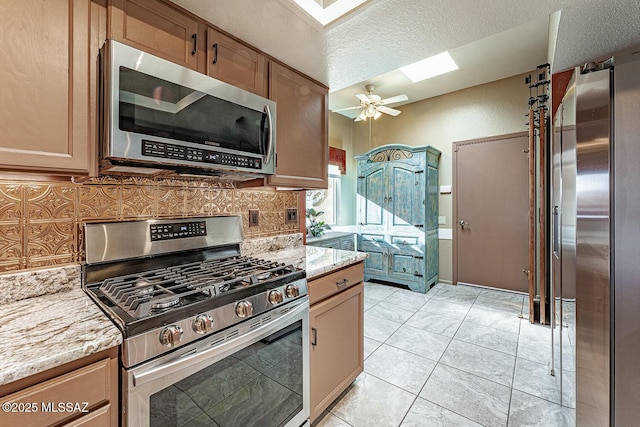 kitchen with a skylight, a ceiling fan, appliances with stainless steel finishes, light stone countertops, and tasteful backsplash