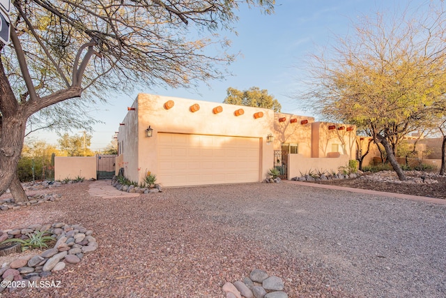 pueblo revival-style home with driveway, an attached garage, a gate, fence, and stucco siding