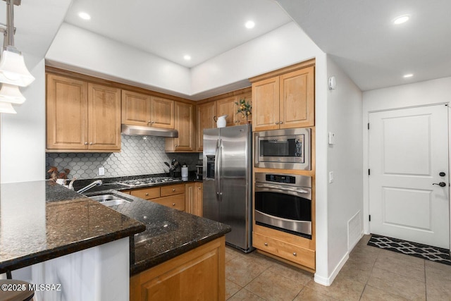 kitchen with under cabinet range hood, a sink, appliances with stainless steel finishes, dark stone counters, and tasteful backsplash