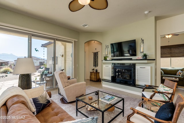 tiled living area featuring arched walkways, a glass covered fireplace, visible vents, and a ceiling fan