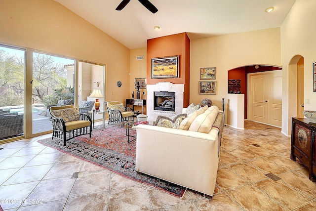 living area with arched walkways, high vaulted ceiling, a tile fireplace, a ceiling fan, and baseboards