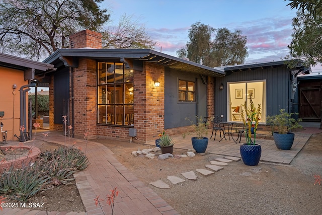 back of house at dusk featuring a chimney, a patio, and brick siding