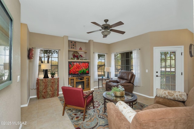 living area featuring light tile patterned floors, baseboards, a ceiling fan, and a glass covered fireplace