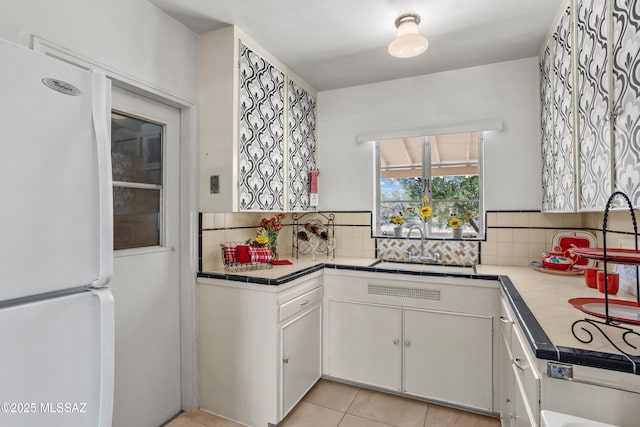 kitchen featuring tile countertops, backsplash, freestanding refrigerator, white cabinetry, and a sink