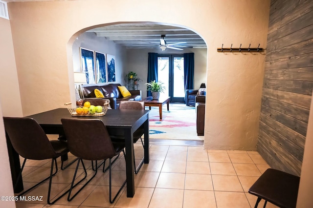 dining space with arched walkways, light tile patterned floors, ceiling fan, wood walls, and beamed ceiling