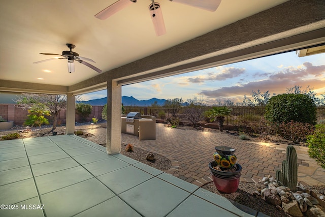 patio terrace at dusk with a fenced backyard, ceiling fan, an outdoor kitchen, and a mountain view