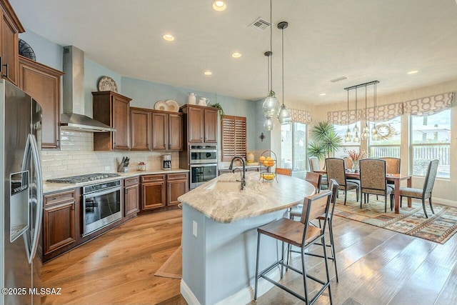 kitchen featuring wall chimney range hood, backsplash, light wood-style flooring, and appliances with stainless steel finishes