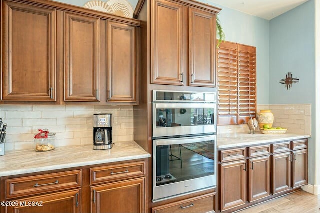 kitchen featuring brown cabinets, light wood-style flooring, decorative backsplash, stainless steel double oven, and light stone countertops