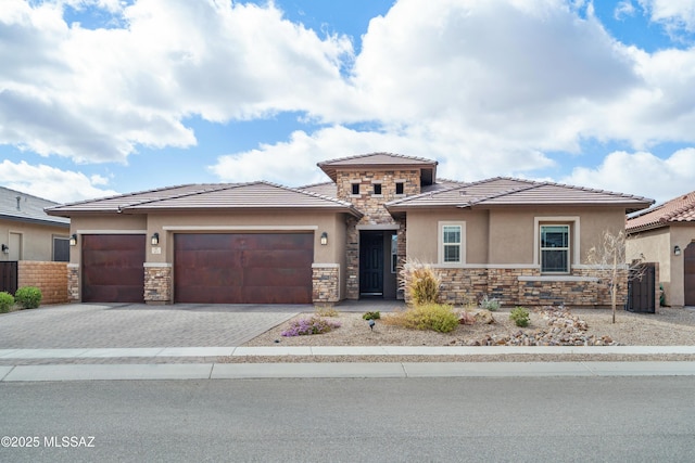 prairie-style house with a garage, fence, stone siding, decorative driveway, and stucco siding