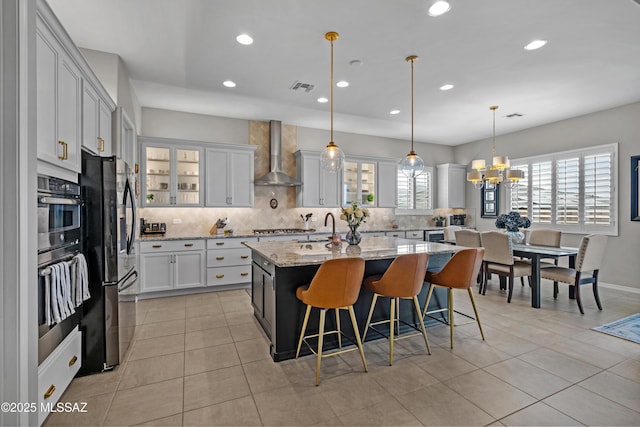 kitchen with tasteful backsplash, appliances with stainless steel finishes, wall chimney range hood, an island with sink, and a kitchen bar