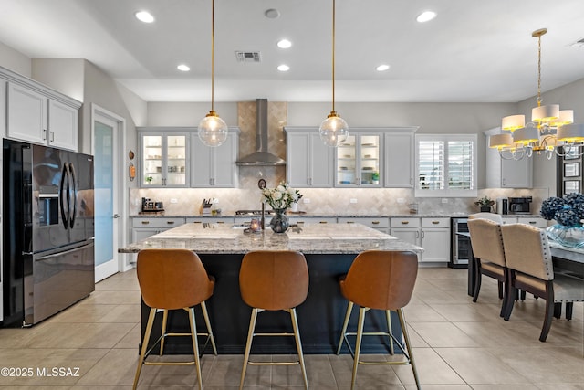 kitchen with black fridge with ice dispenser, visible vents, wall chimney exhaust hood, tasteful backsplash, and an island with sink