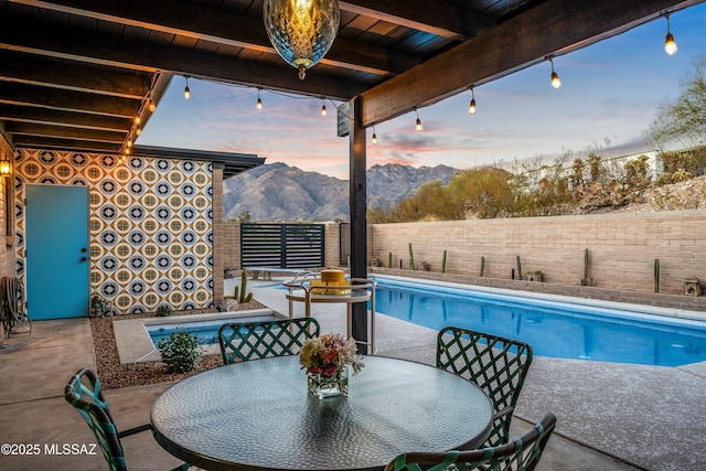 pool at dusk featuring outdoor dining space, a fenced in pool, a fenced backyard, a patio area, and a mountain view