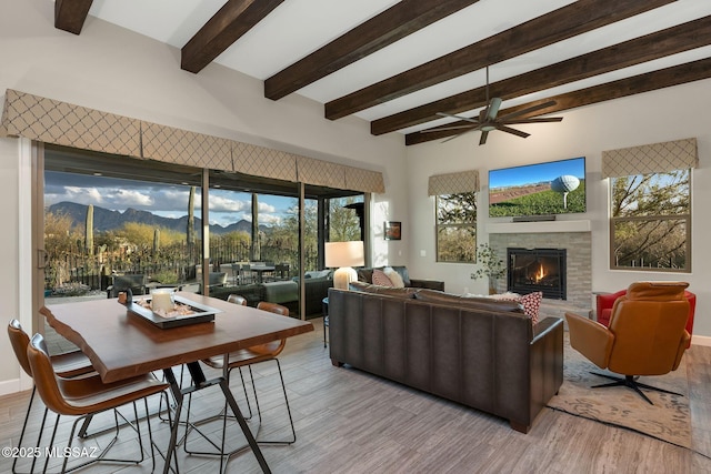 living area with a glass covered fireplace, ceiling fan, light wood-style floors, a mountain view, and beam ceiling