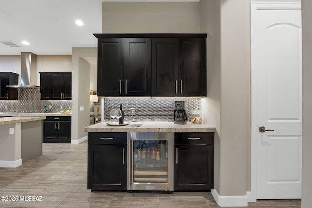 kitchen with beverage cooler, light countertops, wall chimney range hood, and dark cabinets