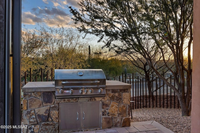 patio terrace at dusk featuring a grill, fence, and an outdoor kitchen