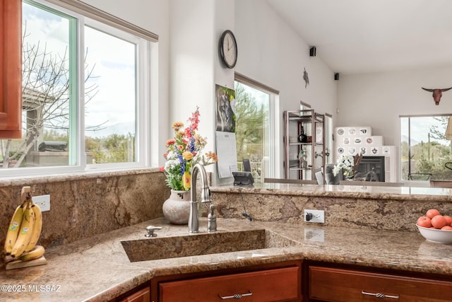 kitchen with light stone counters, brown cabinetry, a sink, and a glass covered fireplace