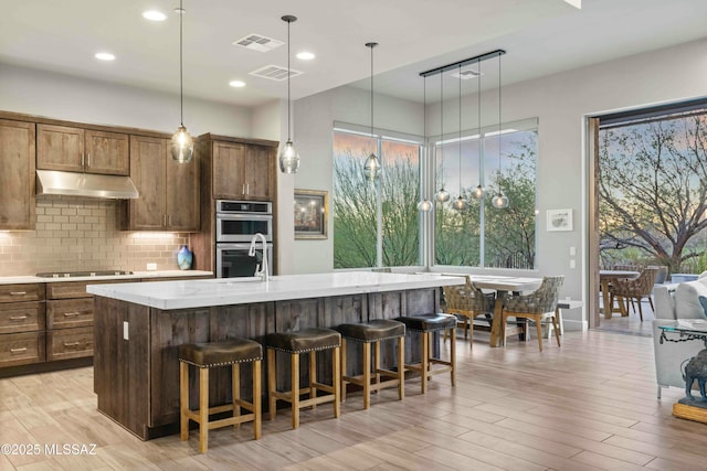 kitchen with double oven, black electric cooktop, under cabinet range hood, visible vents, and decorative backsplash