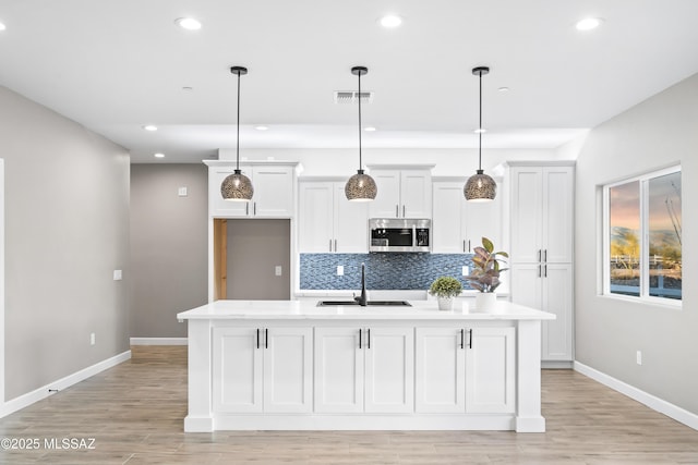 kitchen featuring decorative backsplash, an island with sink, stainless steel microwave, light countertops, and a sink