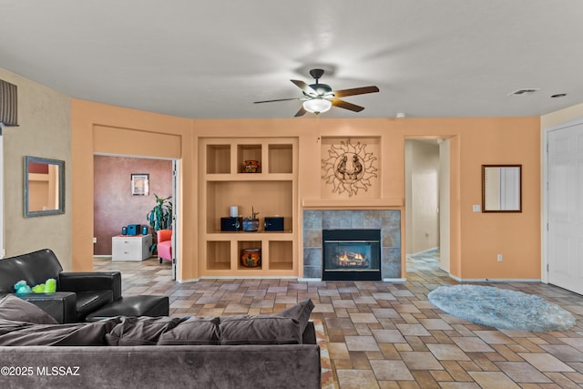 living area with visible vents, a ceiling fan, a tiled fireplace, stone finish floor, and built in shelves