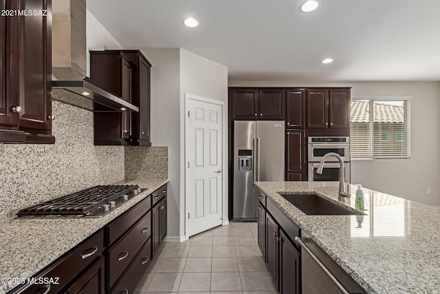 kitchen with decorative backsplash, wall chimney exhaust hood, light stone counters, stainless steel appliances, and a sink