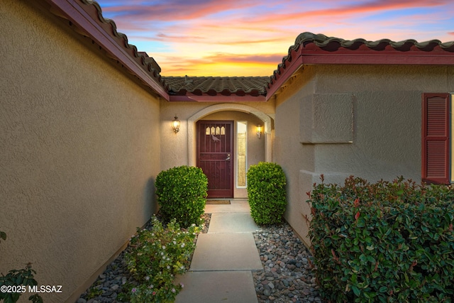 exterior entry at dusk with a tiled roof, an attached garage, and stucco siding