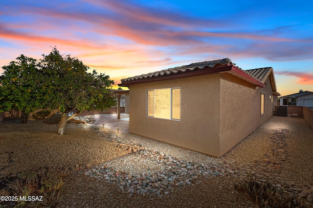 property exterior at dusk with central air condition unit, a tile roof, fence, and stucco siding