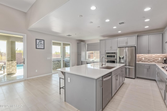 kitchen featuring a kitchen island with sink, gray cabinets, a sink, stainless steel appliances, and decorative backsplash