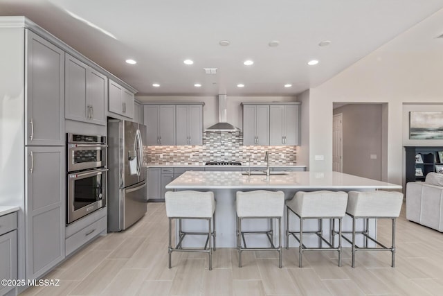 kitchen with wall chimney range hood, a sink, gray cabinetry, appliances with stainless steel finishes, and a kitchen breakfast bar